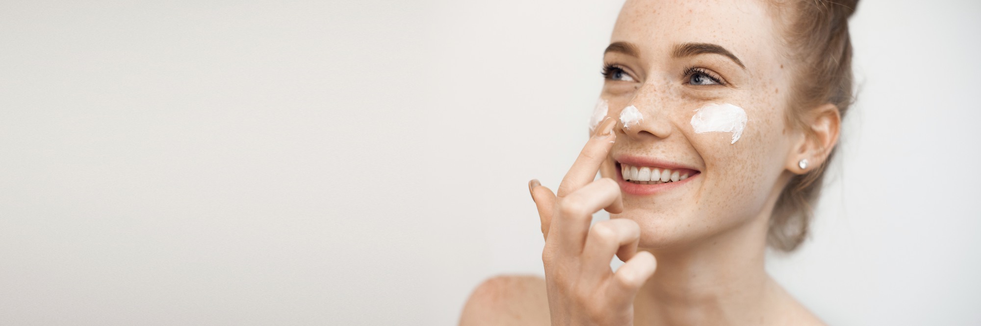 Portrait of a charming young female with red hair and freckles isolated on white applying a anti age cream on her face and nose smiling.